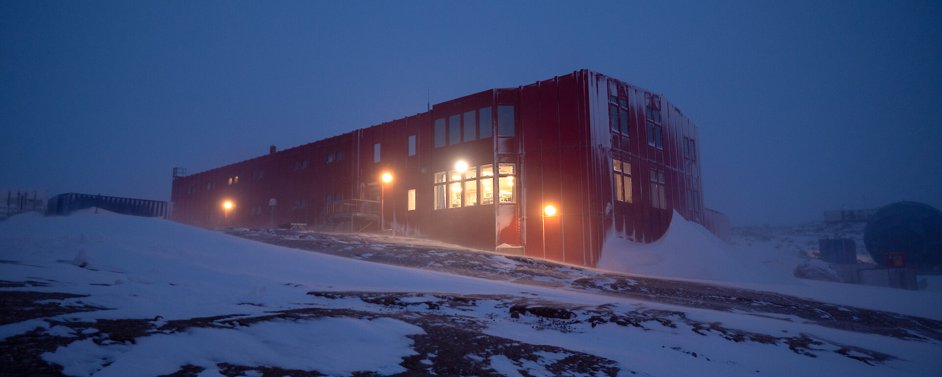 A large red building with lights on stands on a rocky landscape. Snow drifts lie against the building's side.