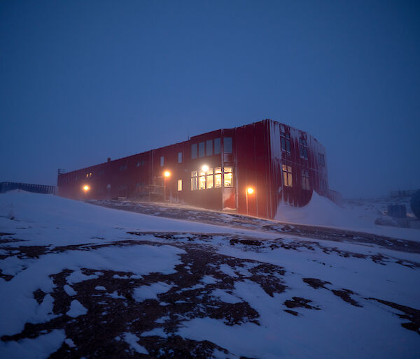 A large red building with lights on stands on a rocky landscape. Snow drifts lie against the building's side.