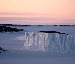 A large iceberg is in the centre of the shot on a frozen sea. There are rocky islands on the left and in the distance.