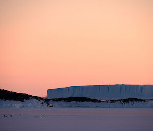 A frozen sea is visible with a very large iceberg in the distance. Three people can just be made out in the lower left of frame on the ice