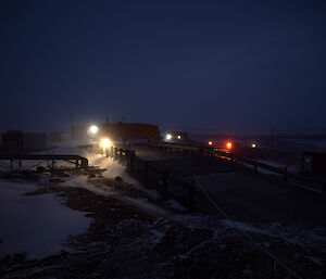 A bridge is seen in the middle of the shot with a large red building in the distance and a dark sky. There are lights on around the building