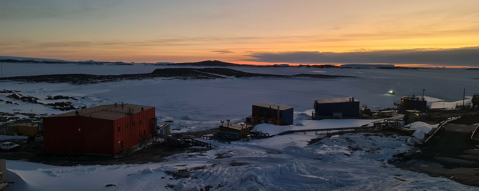The lights of a vehicle can be seen moving over a frozen harbour towards a number of buildings built on a rocky, snow covered landscape.
