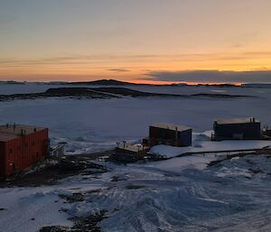 The lights of a vehicle can be seen moving over a frozen harbour towards a number of buildings built on a rocky, snow covered landscape.