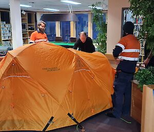 Three men are around a dome tent that has been put up inside in a lounge area. There is a pool table in the background
