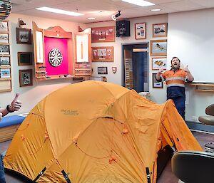 Two men are giving thumbs-up next to a dome tent that has been setup inside in a bar area. There is a darts board and video screen in the background and many photos over the walls