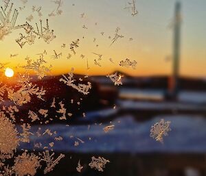 Through a window frosted by ice crystals the sun can be seen just on the horizon. A rocky landscape with some buildings and a tower are blurred in the background.