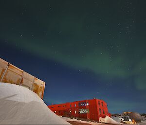 An green aurora is visible in a bright night sky over a large red building. There is a snow drift against an older building in the front left of frame and a satellite dome to the lower right of frame.