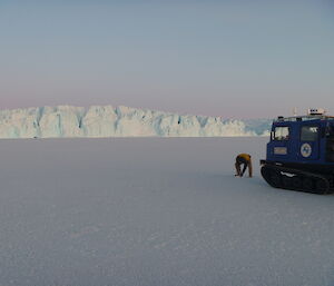 A man is in front of a blue Hägglunds vehicle drilling into the sea-ice. Ice cliffs rise in the distance