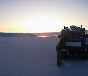 A man is drilling into the sea-ice with a long drill shaft. He is standing in front of a blue Hägglunds vehicle. There is a large iceberg in the distance and the sun is just below the horizon