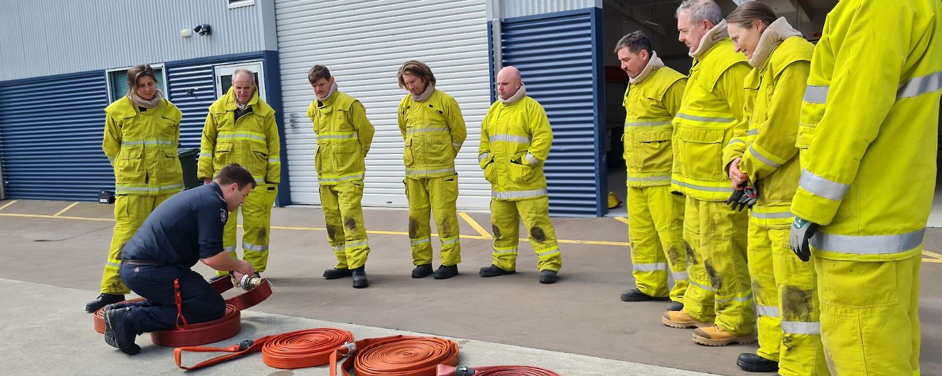 A group of people wearing bright yellow fire-fighting suits watch a man in a blue uniform rolling up a large orange fire hose.
