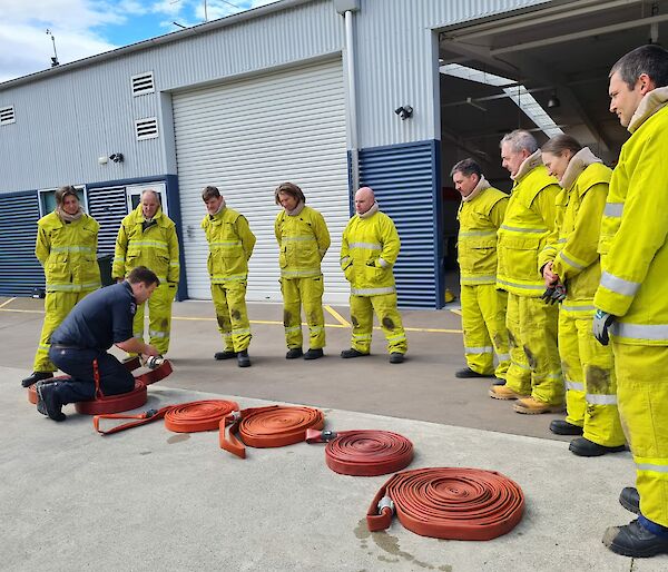 A group of people wearing bright yellow fire-fighting suits watch a man in a blue uniform rolling up a large orange fire hose.