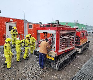 A group of five people wearing yellow fire fighting outfits about to enter a building during a traingin exercise. Another man stands at the back of the fire Hagglunds and is operating the water pump.