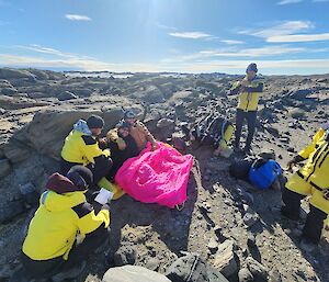 A group of five men rescuing a mock patient who is on the ground in a boulder strewn field. They have covered him in a hot pink bivvy bag to keep him warm.