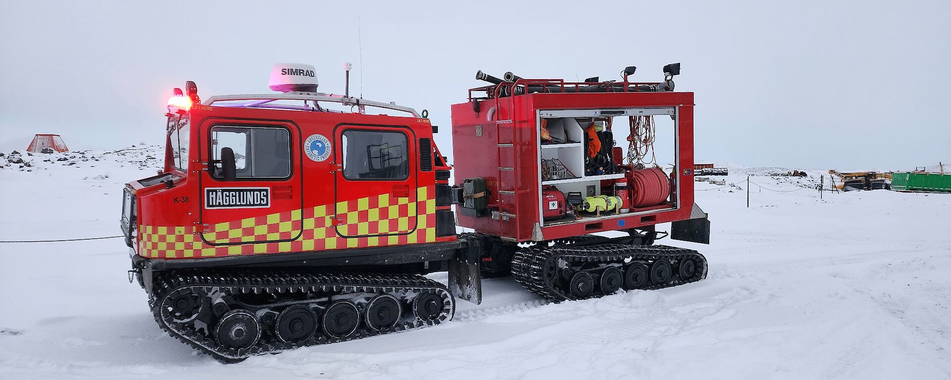 A red Hagglunds vehicle, with a checkerboard stripe in bright yellow down its side, hauls a red trailer containing fire response equipment. The vehicle is parked in the snow.