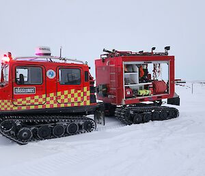 A red Hagglunds vehicle, with a checkerboard stripe in bright yellow down its side, hauls a red trailer containing fire response equipment. The vehicle is parked in the snow.