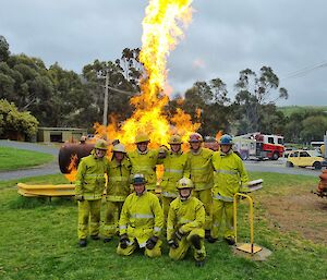 A group of eight smiling people wearing fire-fighters outfits pose for a photograph in front of a huge flame at a fire training centre..