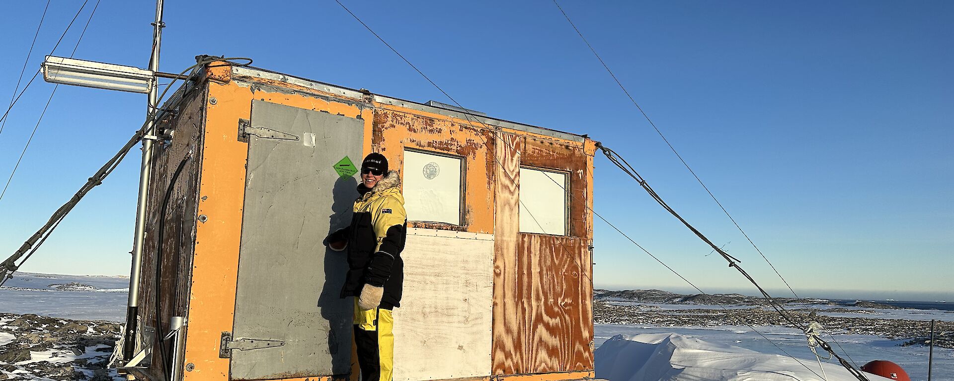 A small wooden hut that stands on a rocky hilltop, elevated on stumps. The hut is anchored to the ground with steel cables and is constructed of bare plywood. A woman stands in front of the closed door, dressed in yellow cold weather clothing and smiling back at the camera.