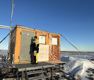 A small wooden hut that stands on a rocky hilltop, elevated on stumps. The hut is anchored to the ground with steel cables and is constructed of bare plywood. A woman stands in front of the closed door, dressed in yellow cold weather clothing and smiling back at the camera.