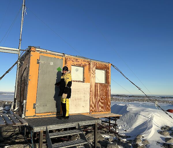 A small wooden hut that stands on a rocky hilltop, elevated on stumps. The hut is anchored to the ground with steel cables and is constructed of bare plywood. A woman stands in front of the closed door, dressed in yellow cold weather clothing and smiling back at the camera.