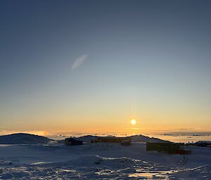 This photo shows a landscape view over rolling, snow covered ground, on towards the horizon and the sun hanging low in the clear blue sky. There are several buildings in the foreground.