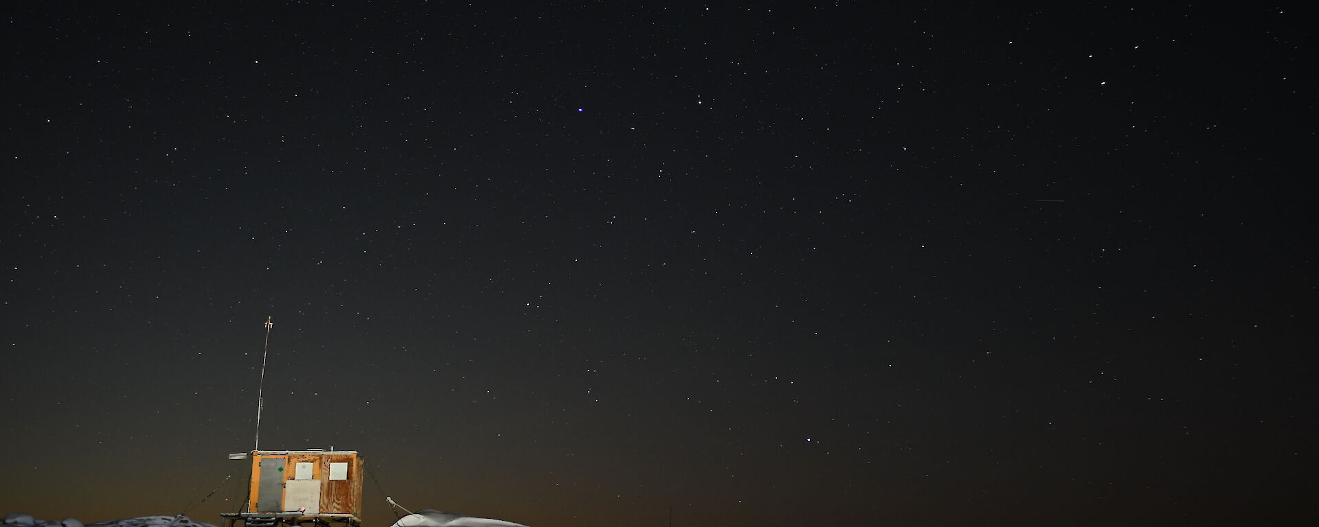 A small wooden hut sits atop a rocky, snow covered hill. It is night time and the sky is filled with stars.