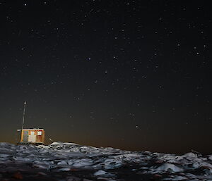 A small wooden hut sits atop a rocky, snow covered hill. It is night time and the sky is filled with stars.