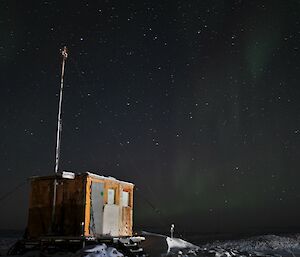 A small wooden hut sits atop a rocky, snow covered hill. It is night time and the sky is filled with stars. There is a faint green and purple aurora active in the sky.