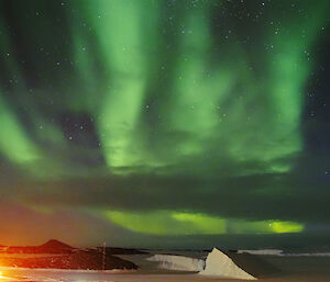 A bright green Aurora is visible in the night sky above icebergs and rocky islands. It appears to make the shape of a hand.
