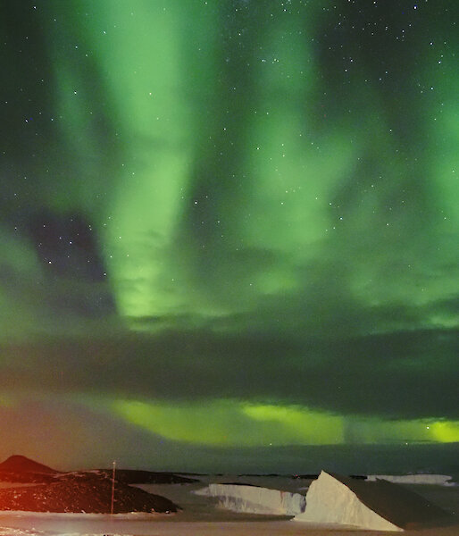 A bright green Aurora is visible in the night sky above icebergs and rocky islands. It appears to make the shape of a hand.