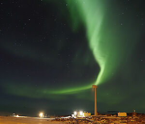 A bright Aurora is in the night sky above a wind turbine and a number of buildings lit by lights.