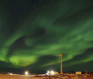 A bright Aurora is in the night sky above a wind turbine and a number of buildings lit by lights.