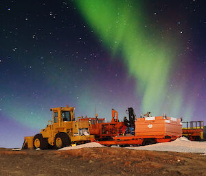 A bright purple and green Aurora is in the night sky above an orange grader and various other trailers and vehicles.