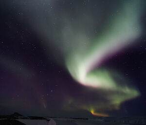 A very bright green, orange, and purple Aurora stretches into the distance in the night sky above an icy landscape