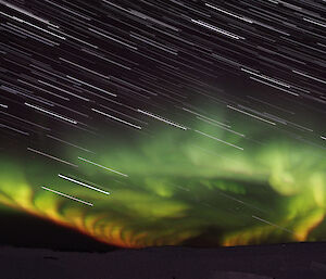 A stacked exposure image showing star trails and a bright green, yellow and orange aurora.