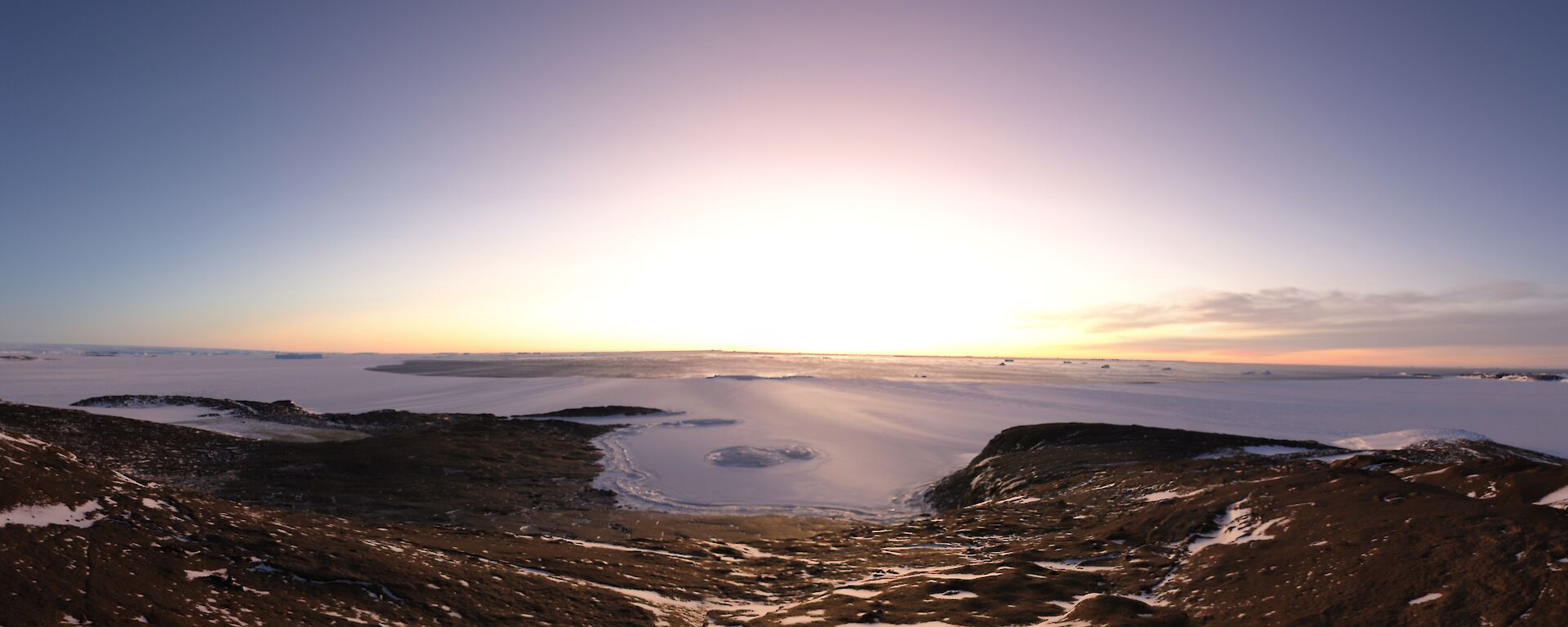 A wide angle view of a ice covered sea beyond a rocky landscape. The sun is setting in the distance over an area of open water