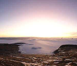 A wide angle view of a ice covered sea beyond a rocky landscape. The sun is setting in the distance over an area of open water