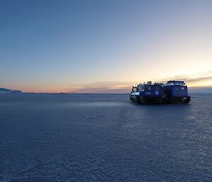 Two Hägglunds vehicles are on the sea-ice with a sunset in the distance to the right of frame and an ice cliff in the left of frame