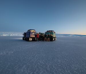 Five men and a woman are standing with two Hägglunds vehicles on the sea-ice. A glacier is in the distance.