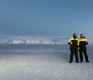 A man and a woman are standing on the sea ice with a glacier in the distance