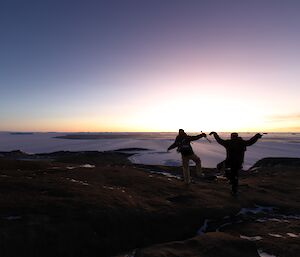 Two men are standing on one foot with their arms in the air on a rocky landscape, a ice covered sea extends into the distance.