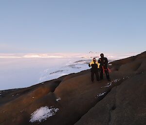 A man and a woman are standing on a rocky hillside with an ice covered sea in the background and ice cliffs and plateau in the far distance