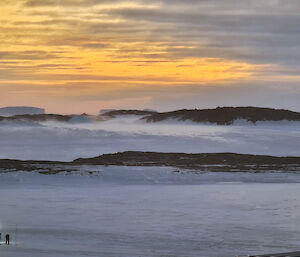 Two men are next to a Hägglunds vehicle in the distance on a ice covered harbour. There is snow blowing over the ice in the far distance towards rocky islands.