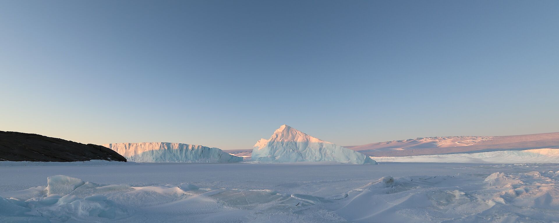 The tips of 2 icebergs reflect the sun. There is a rocky island to the left of frame and an ice-plateau rising in the distance.