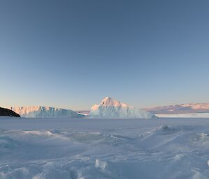 The tips of 2 icebergs reflect the sun. There is a rocky island to the left of frame and an ice-plateau rising in the distance.