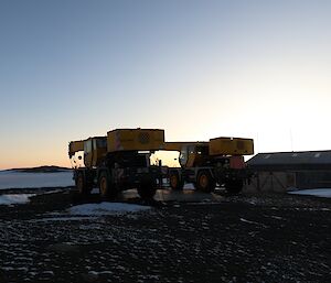 Two large yellow mobile cranes are parked on rocky ground. There is a metal hangar in the background to the right of frame and the glow from the sunset in the distance.
