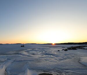 A blue Hägglunds vehicle is driving across a frozen harbour. The sun is setting on the horizon and there is an old metal hangar to the right of frame.