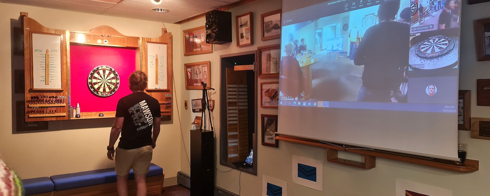 A man is standing near a darts board in a room with many pictures on the wall. There is a projector screen showing people playing darts in another location.