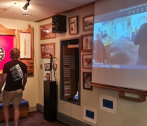 A man is standing near a darts board in a room with many pictures on the wall. There is a projector screen showing people playing darts in another location.