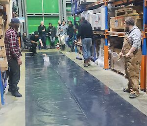 A man is propelling a curling stone (made of ice) across  a strip of black plastic sheeting on the floor of a warehouse.  A number of people are watching on.