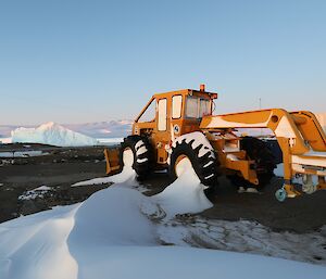 A yellow skidder vehicle with its cab completely filled by snow is on a rocky landscape with two icebergs in the distance.